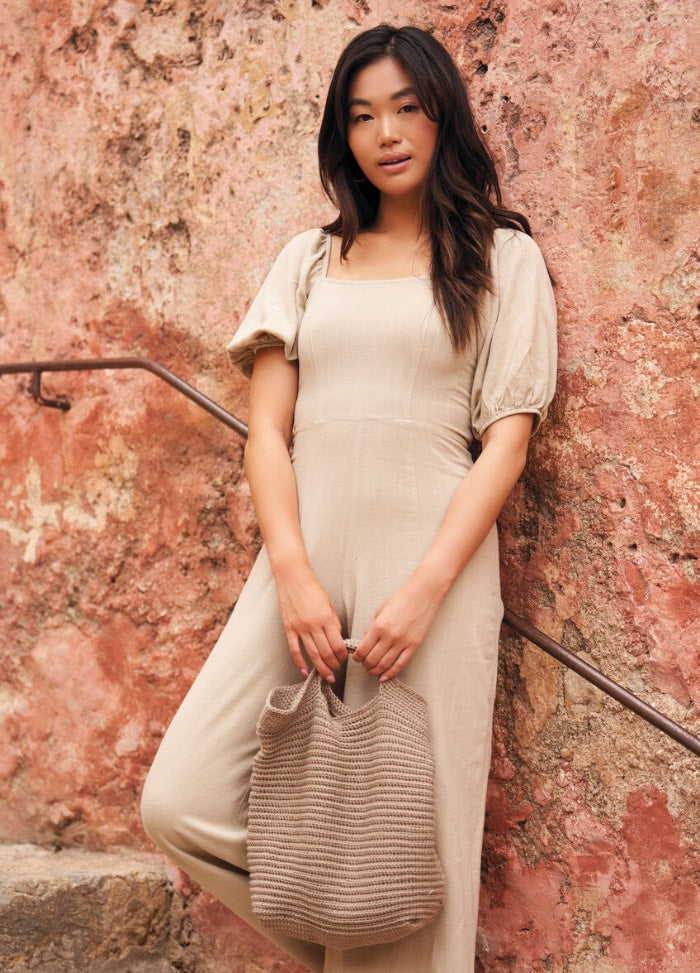 A woman in a beige dress stands against a textured, rustic wall, holding a knitted beige bag. She exudes a relaxed and confident vibe.