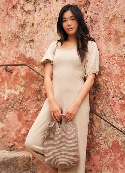 A woman in a beige dress stands against a textured, rustic wall, holding a knitted beige bag. She exudes a relaxed and confident vibe.