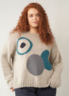 A model with long curly red hair wearing a neutral-toned sweater featuring large abstract circular intarsia shapes in teal, grey, and charcoal against a beige background.