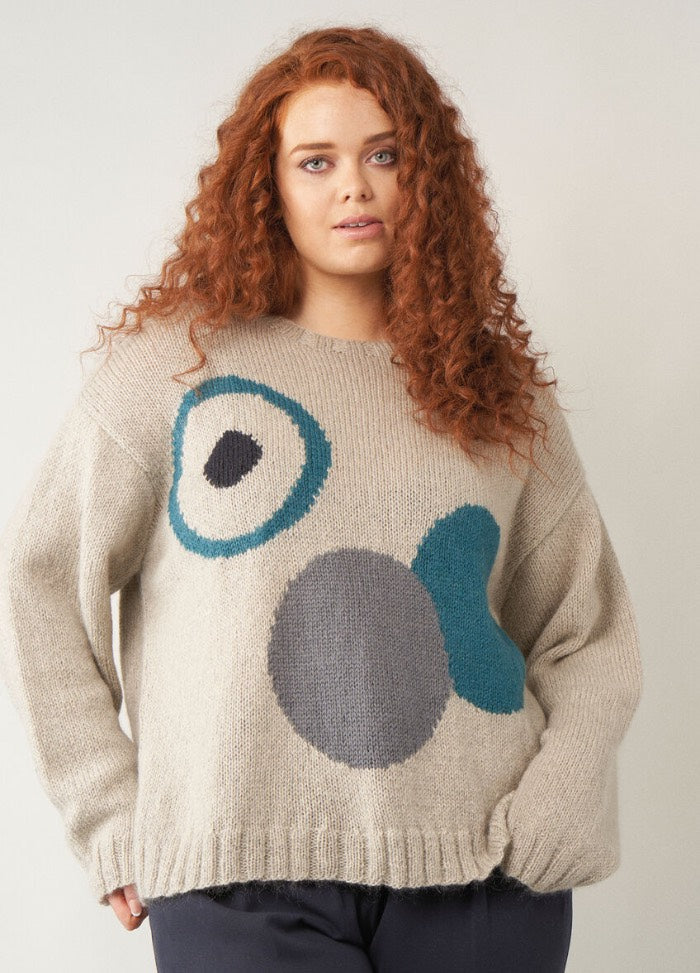 A model with long curly red hair wearing a neutral-toned sweater featuring large abstract circular intarsia shapes in teal, grey, and charcoal against a beige background.