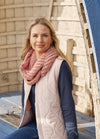 Woman seated on a blue bench, wearing a pink quilted vest and knit scarf over a navy long-sleeve shirt. Background features light wooden structures.