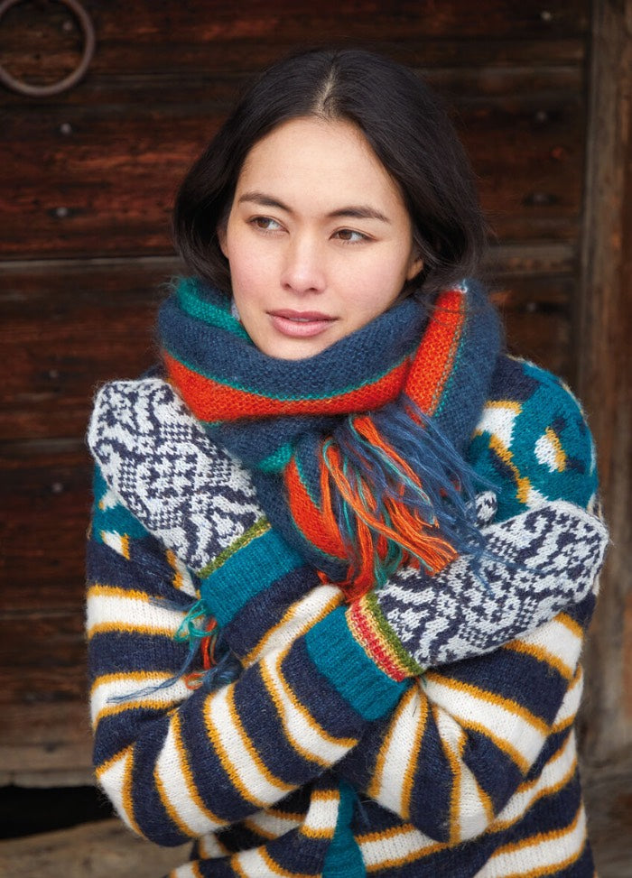 A woman in a colorful striped sweater and patterned mittens, wrapped in a vibrant scarf. She looks thoughtful, standing against a rustic wooden background.