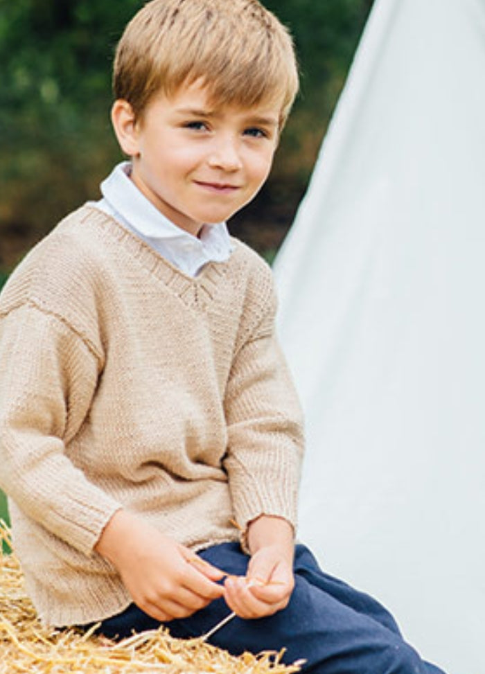 Austin Sweater: Young child sitting on a hay bale, wearing a beige hand-knitted sweater over a white collared shirt and dark pants, with greenery and a white tent in the background.