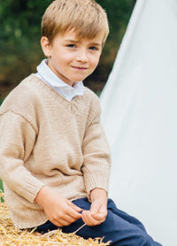 Austin Sweater: Young child sitting on a hay bale, wearing a beige hand-knitted sweater over a white collared shirt and dark pants, with greenery and a white tent in the background.