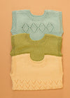 Stack of three hand-knitted baby singlets in soft cotton yarn – top light blue singlet with central diamond motif, middle green singlet in plain stockinette, and bottom yellow singlet featuring a zigzag hem pattern.