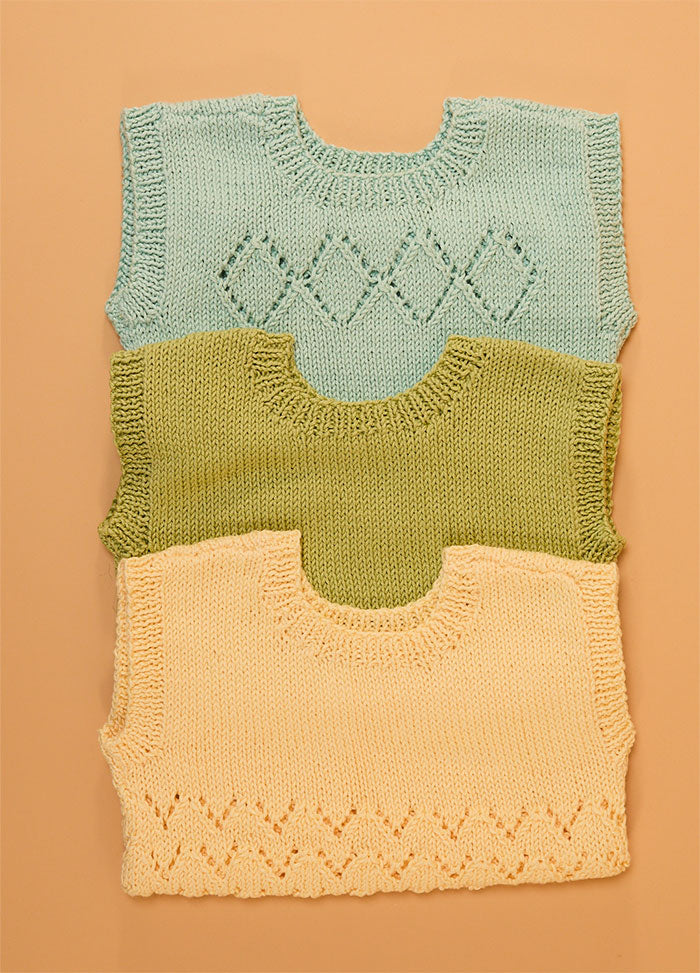 Stack of three hand-knitted baby singlets in soft cotton yarn – top light blue singlet with central diamond motif, middle green singlet in plain stockinette, and bottom yellow singlet featuring a zigzag hem pattern.