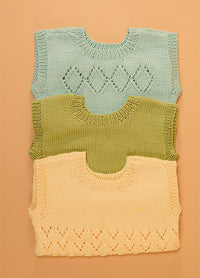 Stack of three hand-knitted baby singlets in soft cotton yarn – top light blue singlet with central diamond motif, middle green singlet in plain stockinette, and bottom yellow singlet featuring a zigzag hem pattern.