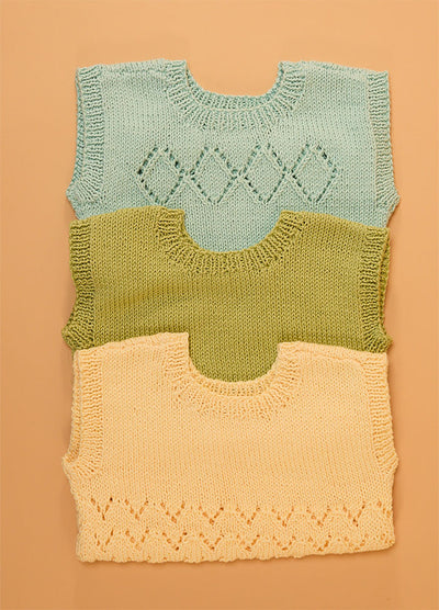 Stack of three hand-knitted baby singlets in soft cotton yarn – top light blue singlet with central diamond motif, middle green singlet in plain stockinette, and bottom yellow singlet featuring a zigzag hem pattern.