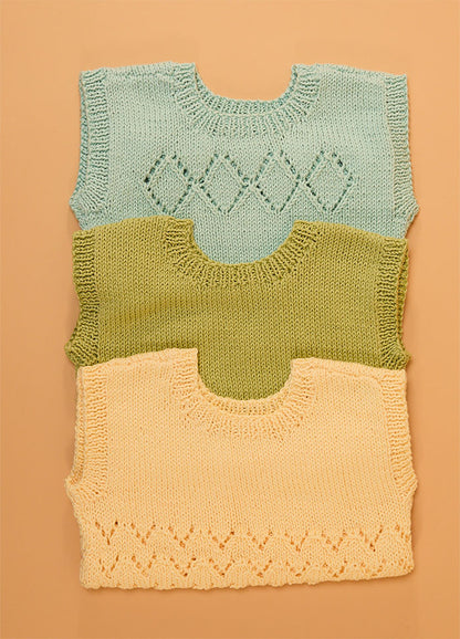 Stack of three hand-knitted baby singlets in soft cotton yarn – top light blue singlet with central diamond motif, middle green singlet in plain stockinette, and bottom yellow singlet featuring a zigzag hem pattern.