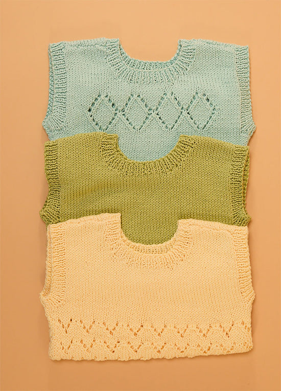 Stack of three hand-knitted baby singlets in soft cotton yarn – top light blue singlet with central diamond motif, middle green singlet in plain stockinette, and bottom yellow singlet featuring a zigzag hem pattern.
