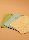 Flat lay of three hand-knitted baby singlets in green, light blue, and cream – each with unique stitch patterns, including a textured diamond design on the cream singlet, displayed on a beige background.