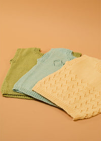 Flat lay of three hand-knitted baby singlets in green, light blue, and cream – each with unique stitch patterns, including a textured diamond design on the cream singlet, displayed on a beige background.