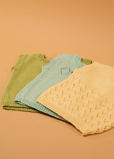 Flat lay of three hand-knitted baby singlets in green, light blue, and cream – each with unique stitch patterns, including a textured diamond design on the cream singlet, displayed on a beige background.
