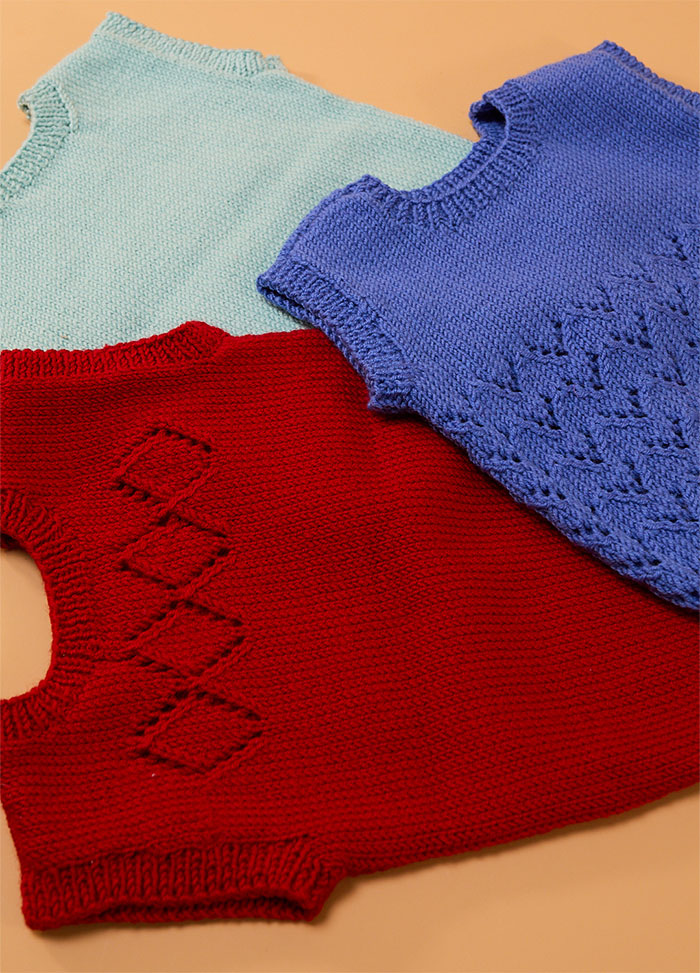 Flat lay of three hand-knitted baby singlets in soft yarn – light blue singlet, dark blue singlet with central diamond motif, and red singlet featuring matching diamond pattern.