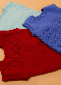 Flat lay of three hand-knitted baby singlets in soft yarn – light blue singlet, dark blue singlet with central diamond motif, and red singlet featuring matching diamond pattern.