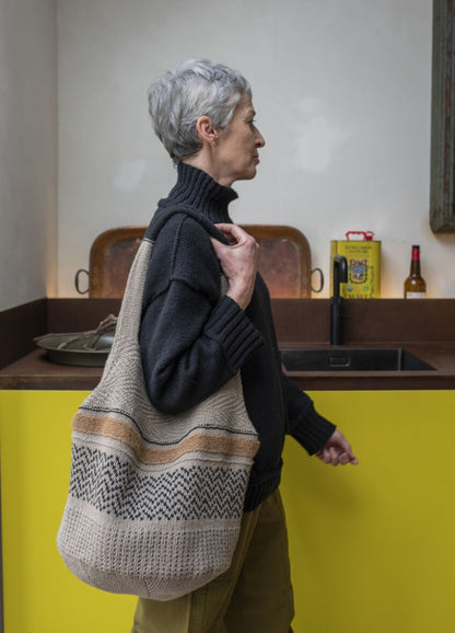 Elderly woman with short gray hair, wearing a dark sweater and carrying a large woven bag, stands in a kitchen with yellow cabinets, creating a cozy atmosphere.