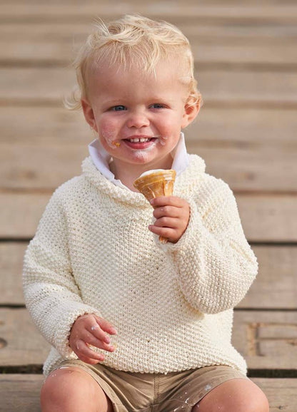 A smiling toddler with messy blonde hair enjoys an ice cream cone, wearing a cozy cream sweater and khaki shorts, seated on wooden steps. Cream smudges on face convey joy.