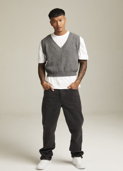 Man in a gray knitted vest over a white t-shirt, wearing black jeans and white sneakers. He stands confidently with hands in pockets against a plain background.