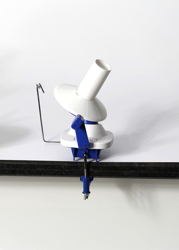 A white and blue hand-operated yarn winder on a table, featuring a clamp for secure use and compact design for easy storage.