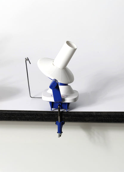 A white and blue hand-operated yarn winder on a table, featuring a clamp for secure use and compact design for easy storage.