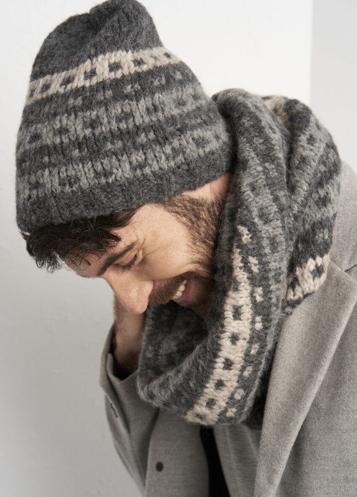 A man in a gray knit hat and scarf ensemble tilts his head while smiling warmly. He wears a light gray coat, conveying a cozy, winter vibe.