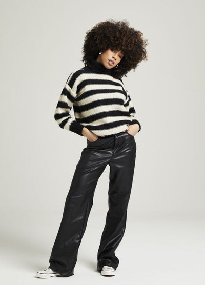 Woman in striped sweater and black leather pants with hands in pockets poses confidently. She has curly hair and wears white sneakers. Neutral background.