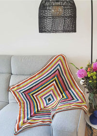 Barcelona Blanket with concentric stripes in pink, yellow, blue, green, and white, draped over a light gray sofa beside a floral arrangement.