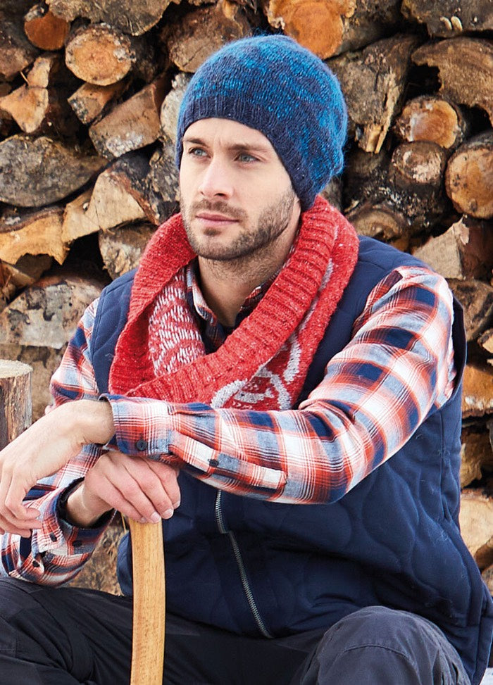 A man in a blue beanie and red scarf sits pensively by a stack of firewood. He wears a plaid shirt and vest, evoking a cozy, autumnal feel.