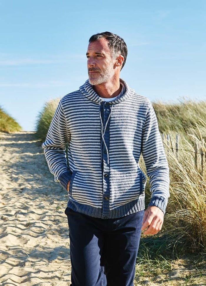 Man in a striped sweater walks on a sandy path flanked by dunes and tall grasses under a clear blue sky, conveying a relaxed, coastal vibe.