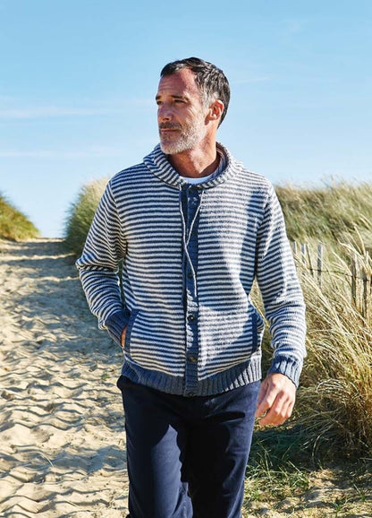 Man in a striped sweater walks on a sandy path flanked by dunes and tall grasses under a clear blue sky, conveying a relaxed, coastal vibe.