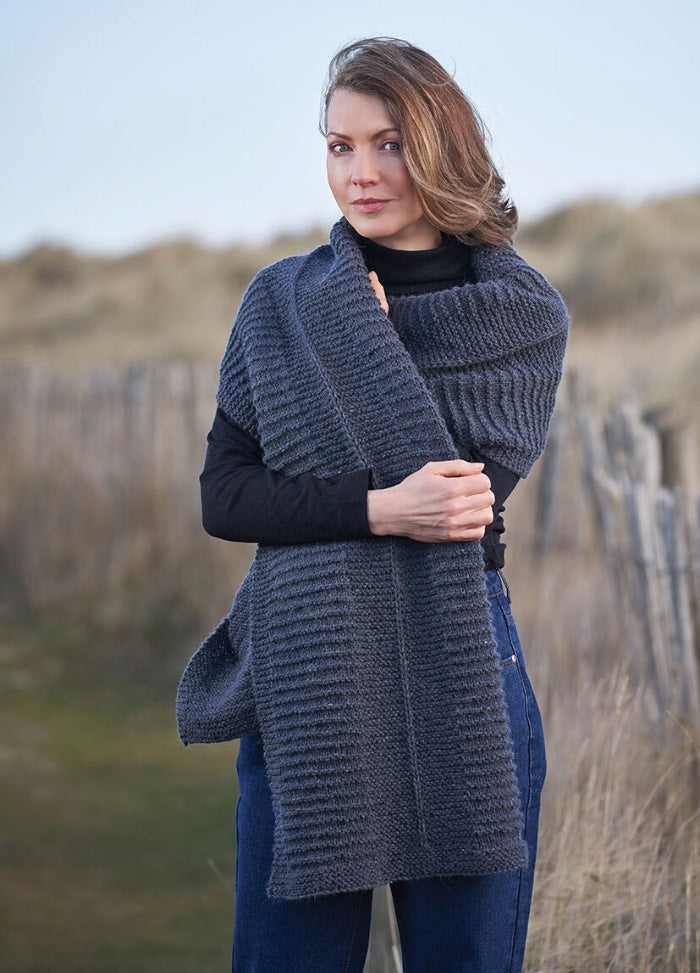 Woman with long hair stands outdoors, wearing a thick gray knitted scarf and blue jeans. The setting is serene with a blurred, natural background.