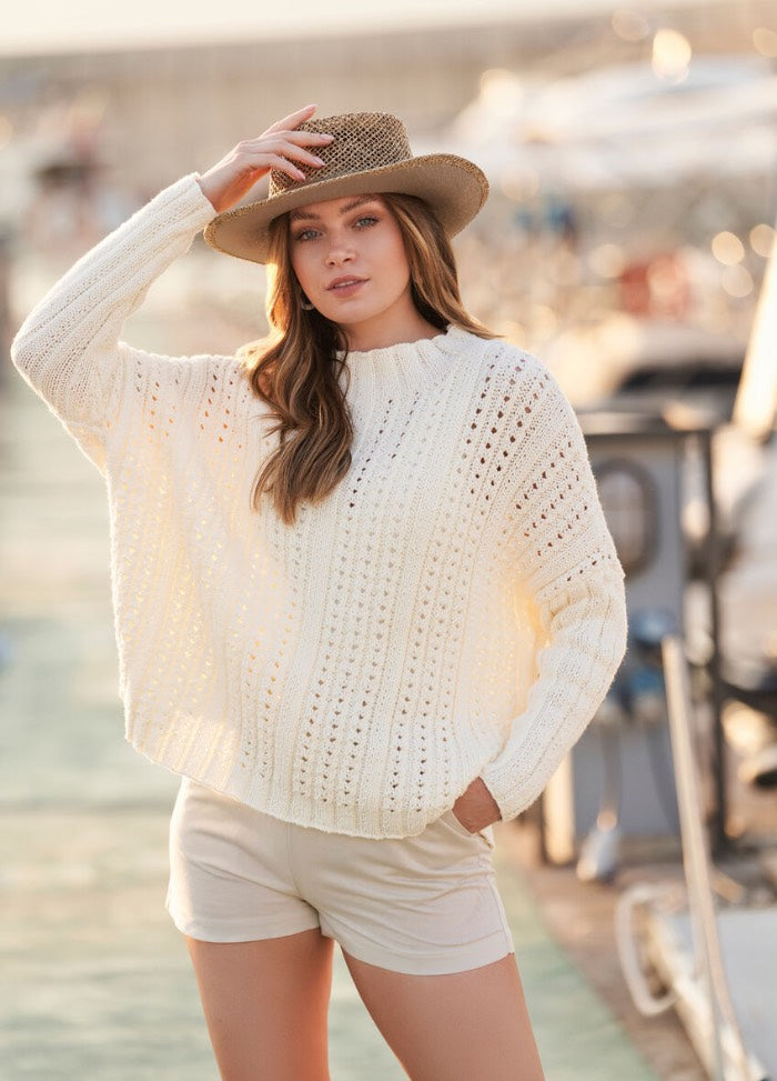 Young woman in a white knit sweater and shorts, wearing a straw hat, stands confidently near a marina. Warm tones and soft lighting evoke a relaxed, summery vibe.