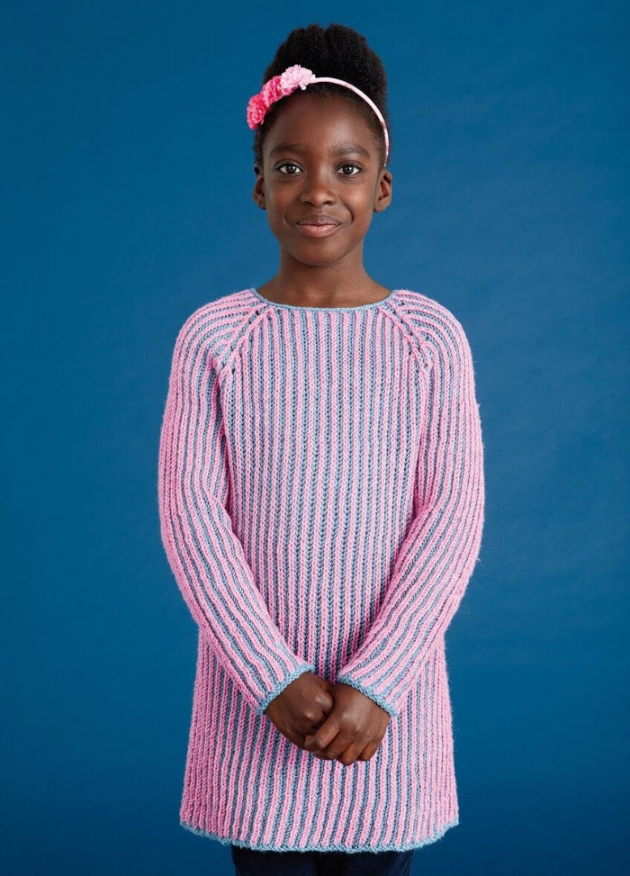 Young girl in a pink and blue striped sweater stands against a blue background, smiling softly. She wears a pink headband, projecting a calm and cheerful vibe.