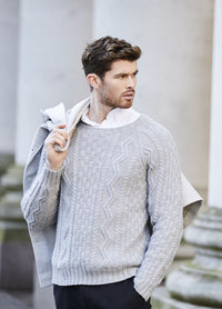 A man with styled brown hair wears a light gray textured sweater over a white shirt. He drapes a jacket over his shoulder, standing outdoors with a confident pose.