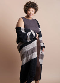 Smiling woman with curly hair wears a sleeveless brown dress and a long, striped cardigan. Neutral background, conveying warmth and comfort.