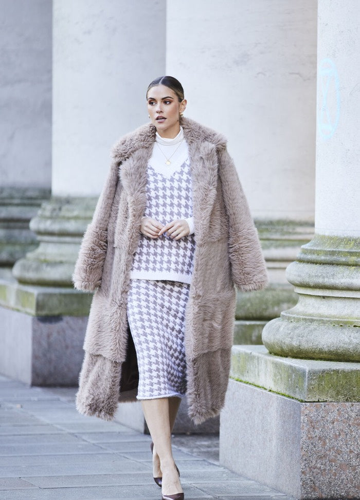 Woman walking confidently in a stylish houndstooth outfit and long, plush beige coat. She passes stone columns, exuding elegance and sophistication.