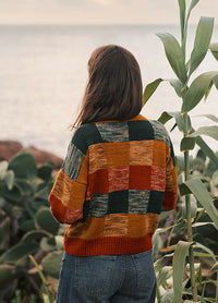Back view of the Berta Sweater, a colorful patchwork knit in green, orange, yellow, and red, worn outdoors near the ocean.