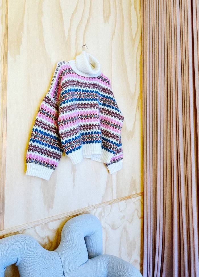 A colourful sweater with patterned stripes of blue, pink, and white hangs on a wooden wall. Below is a light blue, abstract-shaped chair, next to mauve curtains.