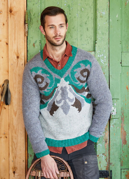 A man with a thoughtful expression leans against a rustic green door. He wears a colorful sweater with teal and brown patterns over a rust shirt.