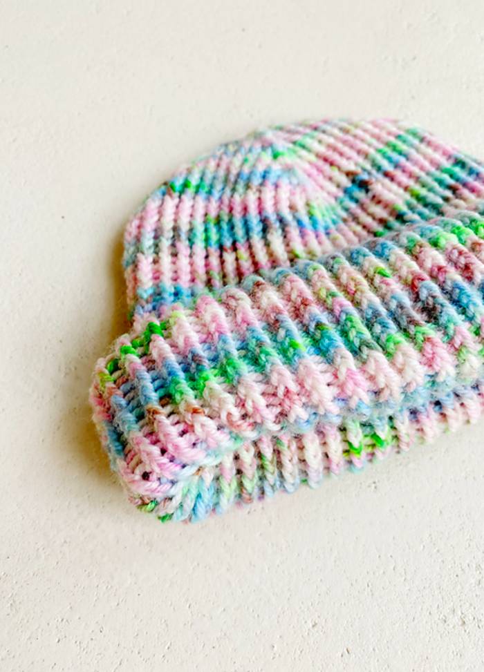 A colourful knitted hat lies on a light surface. It features pastel stripes in pink, blue, green, and white, conveying a cozy, cheerful vibe.