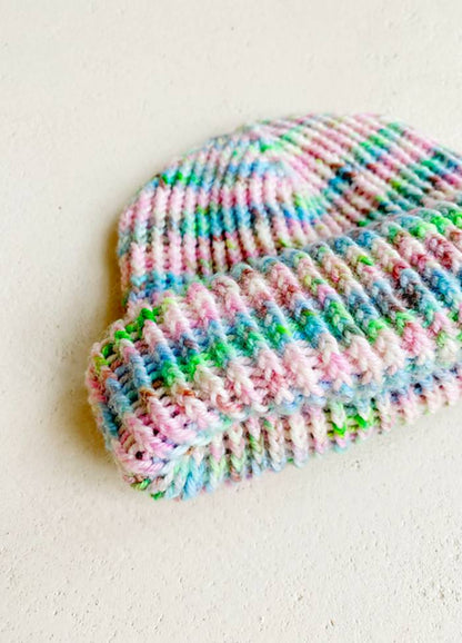 A colourful knitted hat lies on a light surface. It features pastel stripes in pink, blue, green, and white, conveying a cozy, cheerful vibe.