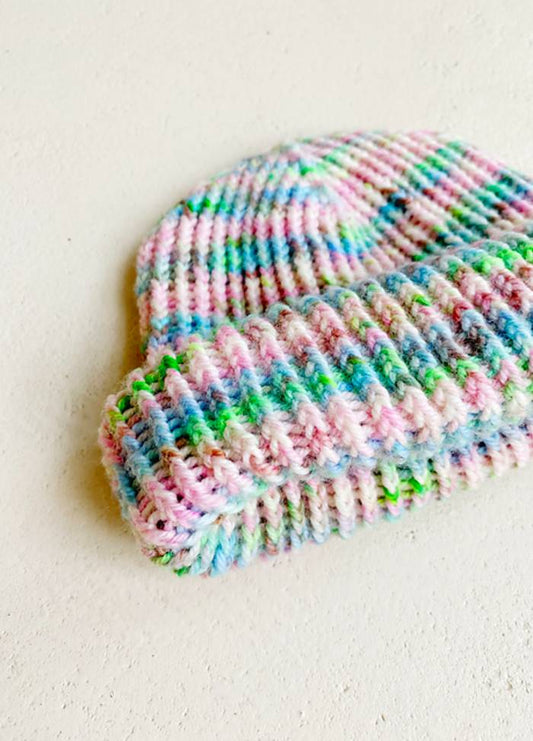 A colourful knitted hat lies on a light surface. It features pastel stripes in pink, blue, green, and white, conveying a cozy, cheerful vibe.