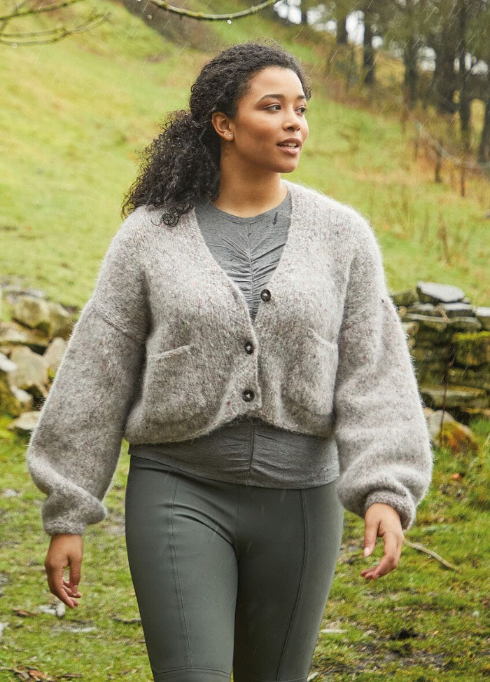 A woman with curly hair walks outdoors, wearing a cozy gray cardigan and leggings. The background features a grassy hillside and stone wall, suggesting a serene, natural setting.