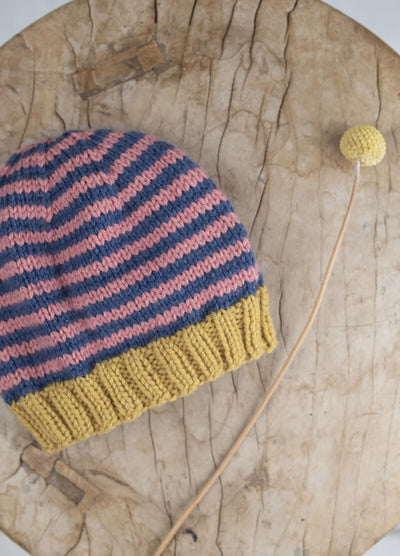Knitted hat with pink and blue stripes, yellow brim on rustic wooden surface. A yellow pom-pom on a stick lies beside, adding a playful touch.