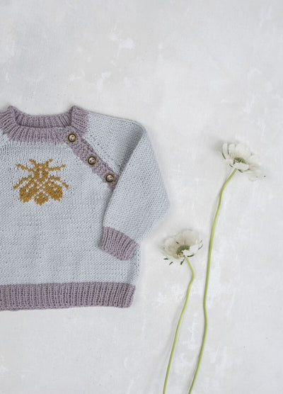 A light blue knitted baby sweater with lavender trim and a yellow bee design lies next to two white flowers on a soft white background. Cozy and delicate.