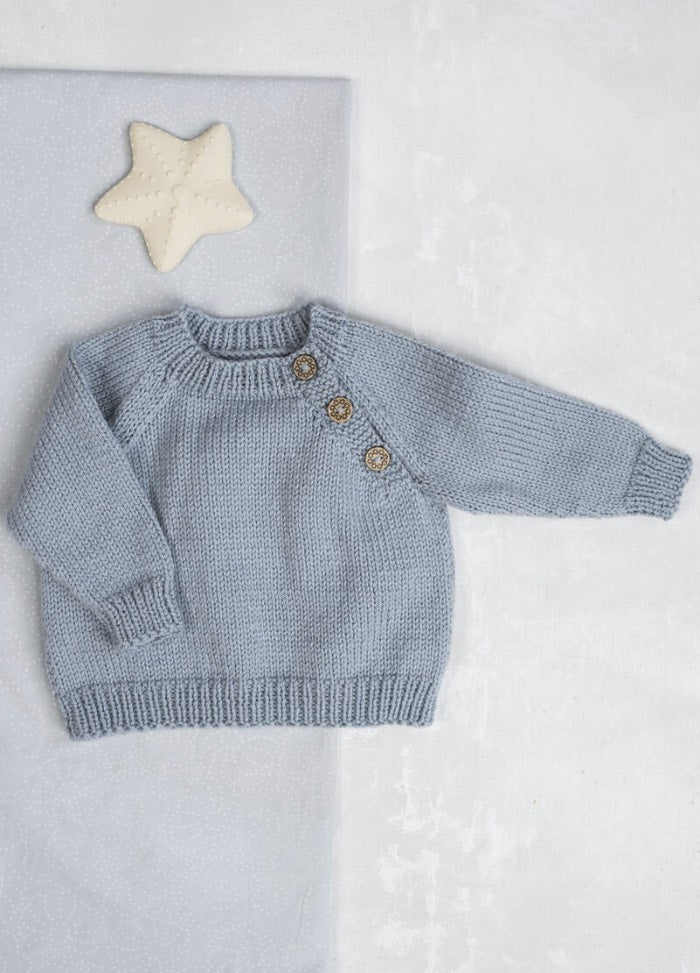 Light blue knitted baby sweater with three buttons on the shoulder, lying on a light surface beside a white star-shaped pillow. Cozy and serene.