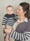 A woman holds a baby wearing matching striped cardigans in gray and navy. She gazes at the baby with warmth and tenderness against a neutral background.
