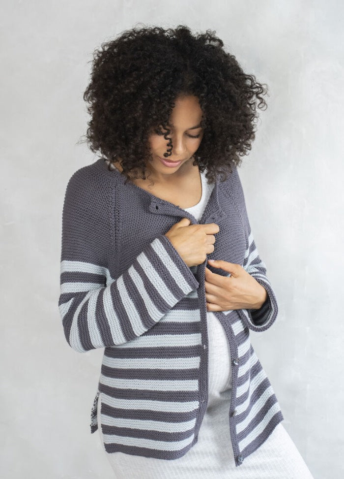 A pregnant woman stands looking down, gently holding her belly. She wears a gray and white striped cardigan over a white dress, conveying a calm, tender mood.