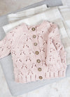 A delicate pink knitted baby cardigan with lace patterns and ornate buttons is neatly displayed on folded, soft, plaid blankets in neutral tones.