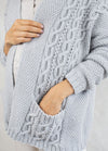 A person wearing a cozy light gray knitted sweater with intricate cable patterns. One hand is in the pocket, conveying warmth and comfort.